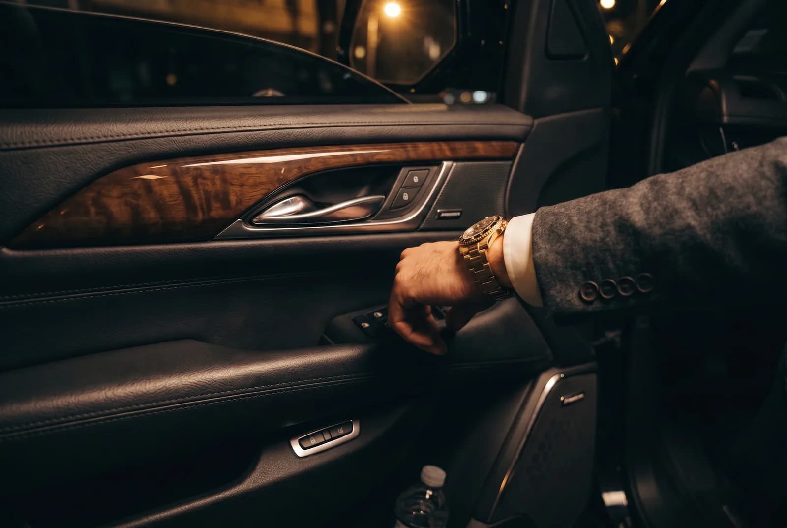 A suited passenger's wrist with a gold watch resting on the car door, burl-wood trim beyond