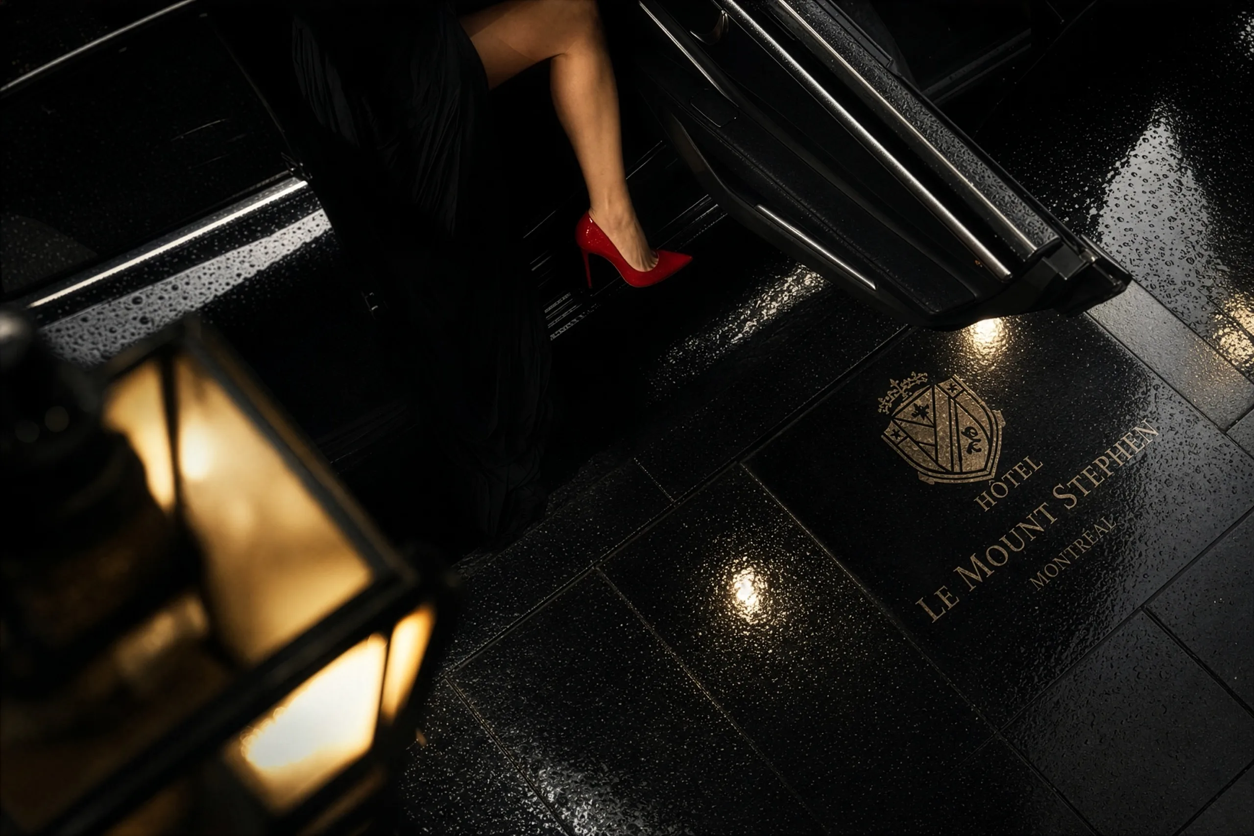 A chauffeur opens the door of a luxury car on a rain-soaked Montréal street at night
